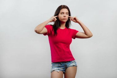 Genç, güzel bir kız onun parmaklarıyla kulaklarını durdurur. Hafif bir arka plan üzerinde izole. Farklı insan duyguları, yüz ifadesi, tutum, algı, beden dili, tepki duyguları.