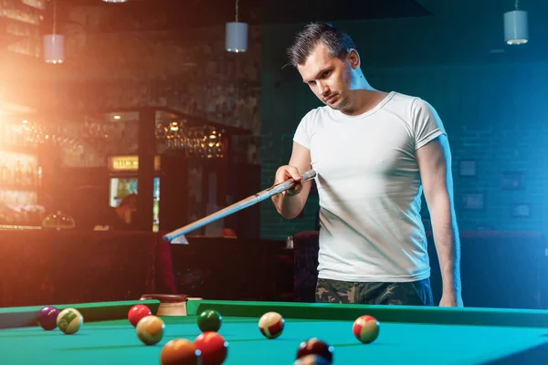 Een man in een wit T-shirt speelt biljart, een donkere achtergrond. Aangenaam tijdverdrijf, entertainment, vrije tijd, gezinsvakanties, spelletjes. — Stockfoto
