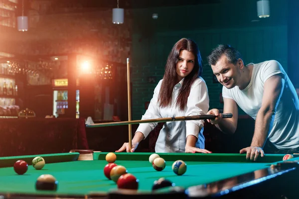A guy and a beautiful girl are playing billiards, a guy is teaching a girl to play billiards. Dark background. Pleasant pastime, family rest, entertainment, holiday.