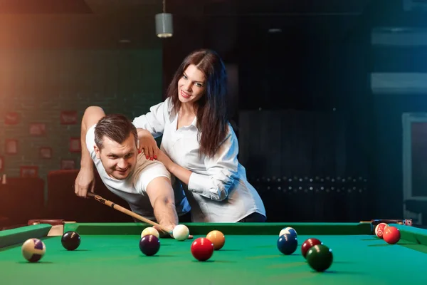 A guy and a beautiful girl are playing billiards, a guy is teaching a girl to play billiards. Dark background. Pleasant pastime, family rest, entertainment, holiday.