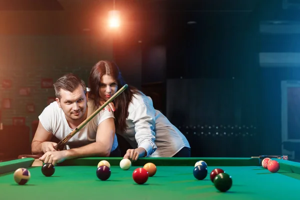 A guy and a beautiful girl are playing billiards, a guy is teaching a girl to play billiards. Dark background. Pleasant pastime, family rest, entertainment, holiday.