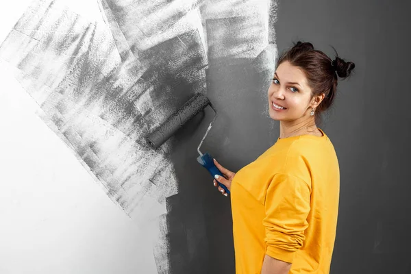 Красивая девушка в желтом свитере рисует стену в серой краске. Покраска комнат, ремонт, дизайн. Копирование пространства . — стоковое фото