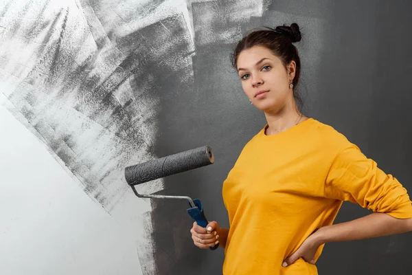 A bela menina em um suéter amarelo pinta uma parede na pintura cinza. Pintura, reparação, design. Espaço de cópia . — Fotografia de Stock