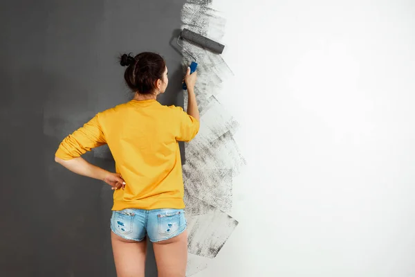 Beautiful girl in a yellow sweater paints a wall in gray paint. Room painting, repair, design. Copy space. — Stock Photo, Image