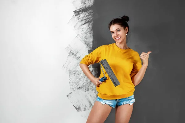 Hermosa chica en un suéter amarillo pinta una pared en pintura gris. Pintura, reparación, diseño. Copiar espacio . — Foto de Stock