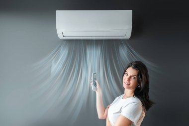 A beautiful girl stands under air conditioning and a stream of fresh cold air against a gray wall. The concept of heat, cool air, cooling, freshness. clipart