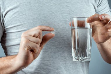 Male hands close-up, holds a glass of water and a pill, medication, treatment, gray background. Medical theme, vitamins, health care.