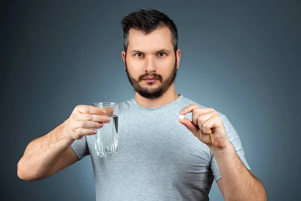 Un homme tient un verre d'eau et une pilule, prenant des médicaments, un traitement, fond gris. Thème médical, vitamines, soins de santé . — Photo