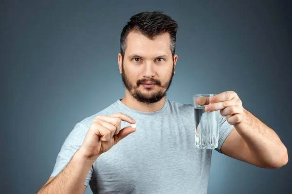 Un homme tient un verre d'eau et une pilule, prenant des médicaments, un traitement, fond gris. Thème médical, vitamines, soins de santé . — Photo