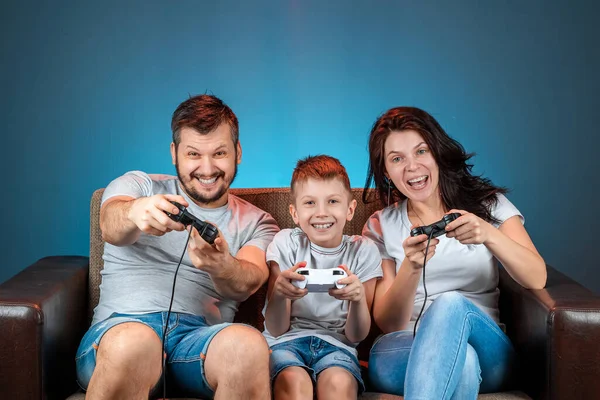 A cheerful family, dad mom and son play on the console, video games, emotionally react sitting on the couch. Day off, entertainment, leisure, spend time together.