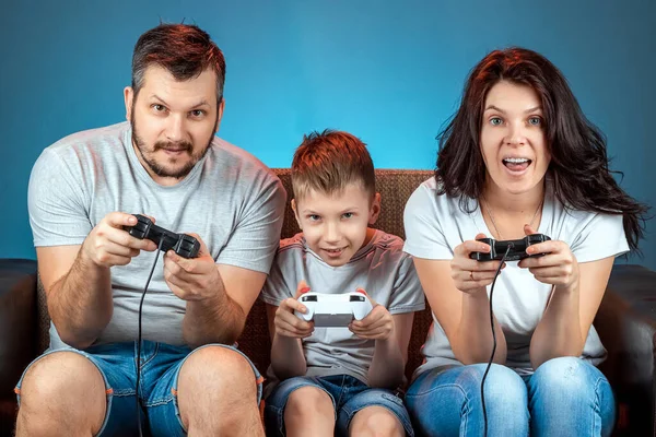 A cheerful family, dad mom and son play on the console, video games, emotionally react sitting on the couch. Day off, entertainment, leisure, spend time together.