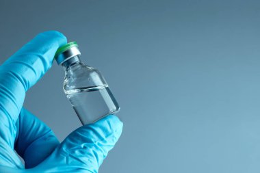 Hand in a blue glove holds a bottle with a vaccine close-up on a gray background, medical concept vaccination subcutaneous injection, dose. Disease treatment immunization.