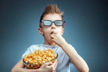 The boy holds in his hands and eats popcorn watching a movie in 3D glasses, blue background. The concept of a cinema, films, emotions, surprise, leisure. streaming platforms.