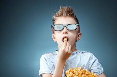 The boy holds in his hands and eats popcorn watching a movie in 3D glasses, blue background. The concept of a cinema, films, emotions, surprise, leisure. streaming platforms.