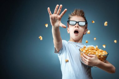 A boy holding popcorn in his hands watching a movie in 3D glasses, fear, blue background. The concept of a cinema, films, emotions, surprise, leisure. streaming platforms.