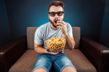 A man watches a movie or series with 3D glasses, a blue background. The concept of a cinema, films, emotions, surprise, leisure, streaming platforms.