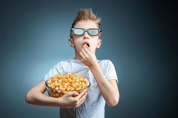 Le garçon tient dans ses mains et mange du pop-corn en regardant un film en lunettes 3D, fond bleu. Le concept de cinéma, films, émotions, surprise, loisirs. plateformes de streaming . — Photo