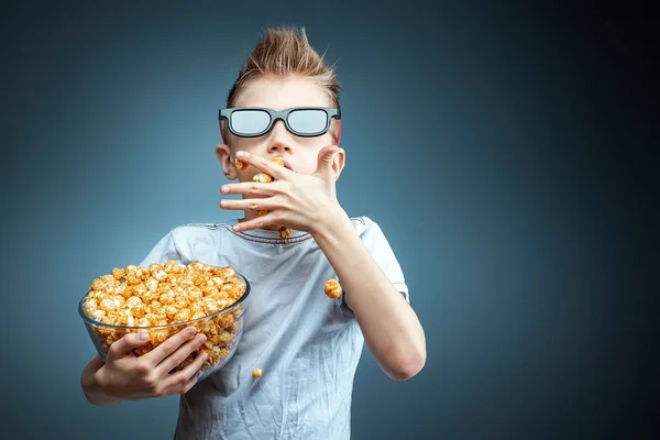 Hält der Junge in den Händen und isst Popcorn, während er einen Film in 3D-Gläsern anschaut, blauer Hintergrund. das Konzept eines Kinos, Filme, Emotionen, Überraschung, Freizeit. Streaming-Plattformen. — Stockfoto