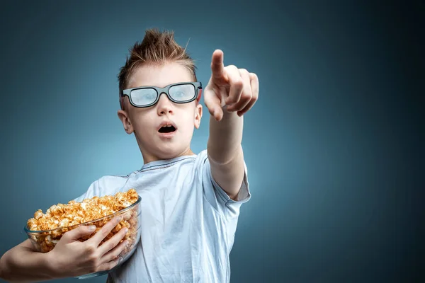 The boy holds popcorn in his hands watching a movie in 3D glasses on a blue background. The concept of a cinema, films, emotions, surprise, leisure. — 图库照片