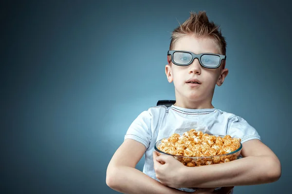 Le garçon tient du pop-corn dans ses mains en regardant un film en lunettes 3D sur un fond bleu. Le concept de cinéma, films, émotions, surprise, loisirs . — Photo