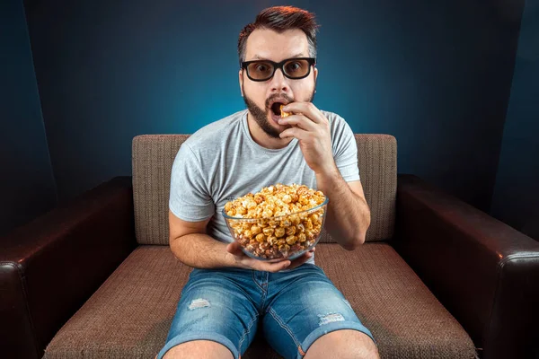 Un hombre ve una película o serie con gafas 3D, un fondo azul. El concepto de un cine, películas, emociones, sorpresa, ocio, plataformas de streaming . —  Fotos de Stock