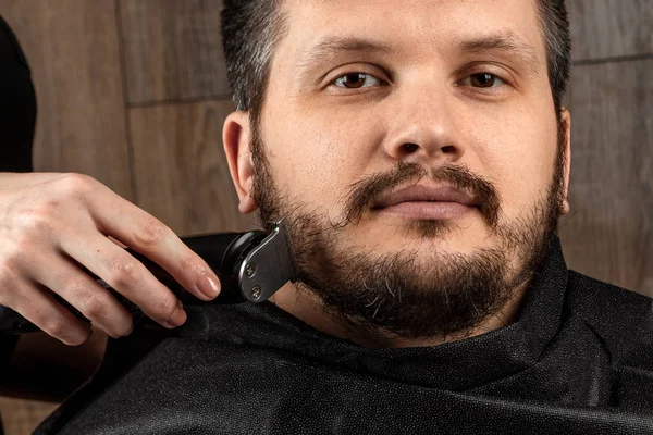 The master makes a beard haircut to the client with an electric machine, trimmer, close-up. The process of shaving a beard at the hairdresser. Body care, lifestyle, metrosexual.