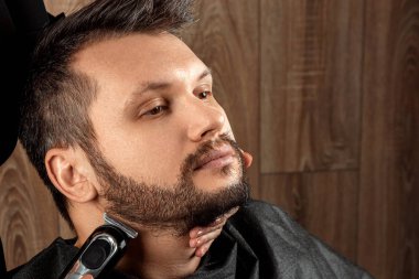 Usta, elektrikli makina ile müşteriye sakal tıraşı yapar, budayıcı, yakın plan. Kuaförde sakal kesme işlemi. Vücut bakımı, yaşam tarzı, metroseksüel..