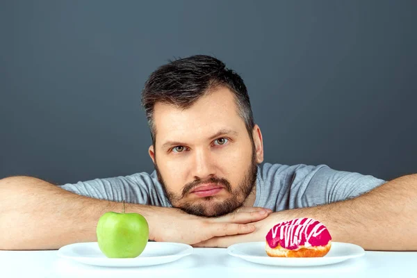 Ein erwachsener Mann hat die Wahl zwischen einem Donut und einem grünen Apfel. Konzept Widerstand gegen Versuchung, Fast Food, gesunde Ernährung, Ernährung, Körperpflege. — Stockfoto