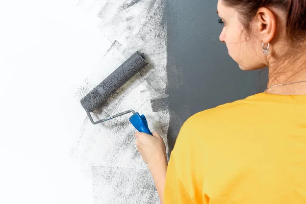 Menina bonita pinta uma parede em uma jaqueta amarela com um rolo, tinta cinza. O conceito de reparação, mudança, design, interior . — Fotografia de Stock