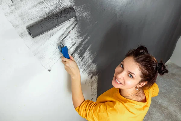 Menina bonita pinta uma parede em uma jaqueta amarela com um rolo, tinta cinza. O conceito de reparação, mudança, design, interior . — Fotografia de Stock
