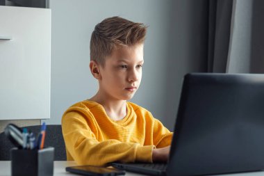 Sarı ceketli bir çocuk, evde bir masada oturuyor, bir dizüstü bilgisayara bakıyor, internetten öğreniyor, evde internet üzerinden öğreniyor. Teknoloji, okul, bilgi. Boşluğu kopyala