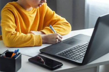 Kalemle yakın plan ödev yapan bir çocuğun eli. Çevrimiçi öğrenme kavramı, evde uzaktan öğrenme, teknoloji, okul. Yumuşak odak