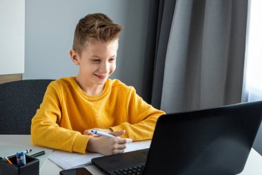 Sarı ceketli bir çocuk, evde bir masada oturuyor, bir dizüstü bilgisayara bakıyor, internetten öğreniyor, evde internet üzerinden öğreniyor. Teknoloji, okul, bilgi. Boşluğu kopyala