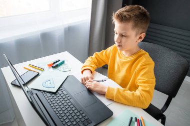 Sarı ceketli bir çocuk, evde bir masada oturuyor, bir dizüstü bilgisayara bakıyor, internetten öğreniyor, evde internet üzerinden öğreniyor. Teknoloji, okul, bilgi. Boşluğu kopyala