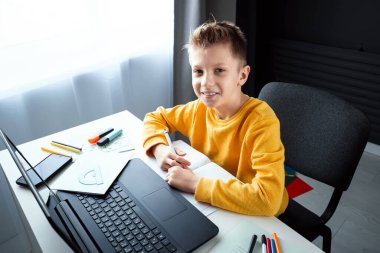 Sarı ceketli bir çocuk, evde bir masada oturuyor, bir dizüstü bilgisayara bakıyor, internetten öğreniyor, evde internet üzerinden öğreniyor. Teknoloji, okul, bilgi. Boşluğu kopyala