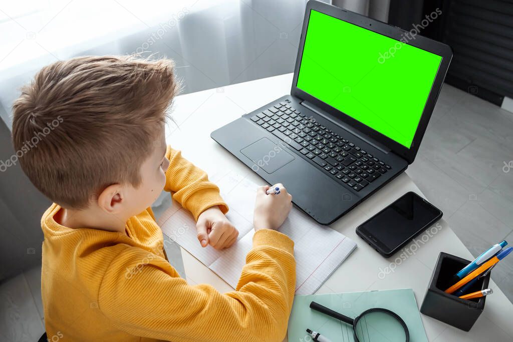 A child, a boy in a yellow sweater, is sitting at a table at home looking in a beech laptop with a green screen. The concept of online learning, distance learning at home, technology, school