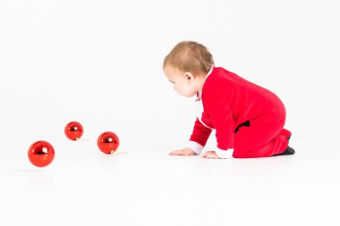 Stüdyo fotoğrafı, beyaz arka planda Noel Baba kılığında Noel toplarıyla oynayan bir bebek.