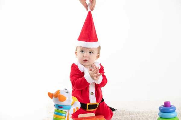 Bebê disfarçado de Papai Noel sentado no chão rolante de brinquedos, enquanto uma mão remove o chapéu de sua cabeça — Fotografia de Stock