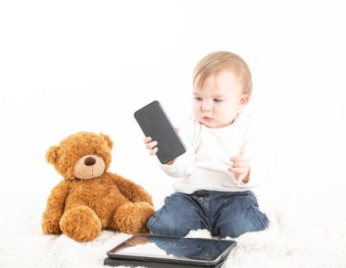 Elinde cep telefonu ve yerde oyuncak ayısı olan bir bebek.