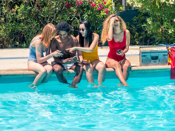 Stock photo of group of people of different ethnic groups with bathing suits of different colors with the mobile in the hand sitting on the edge of a swimming pool with a cassette and a lgtb flag. Lifestyle