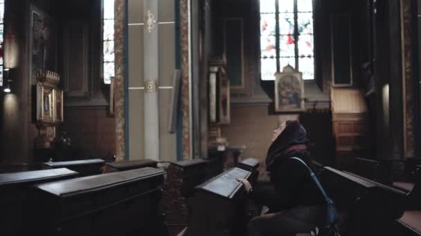 Молодая женщина сидит в католической церкви. Девушка в кафтане: внутри святого места — стоковое видео
