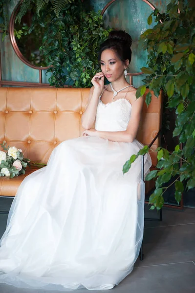 Aziatische bruid met bloemen in witte jurk zitten op de bank — Stockfoto