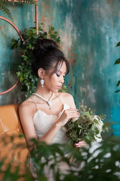 Asian bride with flowers in white dress sitting on sofa