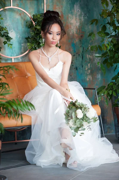 Asian bride with flowers in white dress sitting on sofa — ストック写真