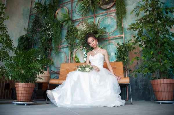 Asian bride with flowers in white dress sitting on sofa — Stock Photo, Image