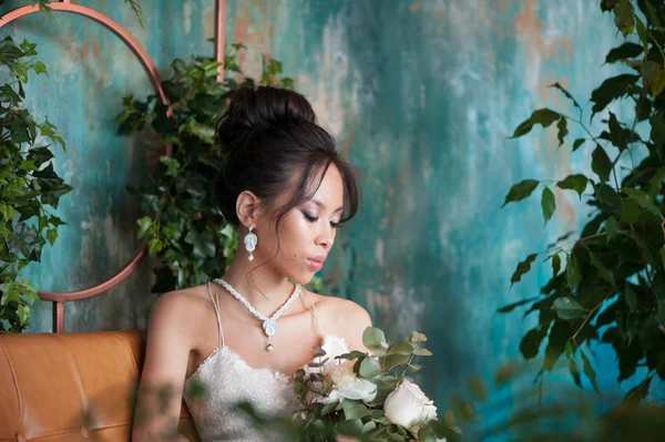 Азиатская невеста с цветами в белом платье сидит на диване — стоковое фото