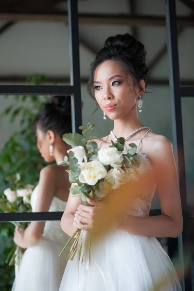 Asian bride with flowers in white dress. Indoor room back to mirror