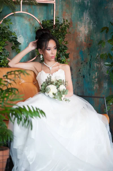 Asian bride with flowers in white dress sitting on sofa indoor floral room