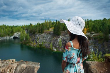 Elbiseli kadın arkası ve güzel manzaranın arkasında beyaz hasır şapka, göl ve bulutlu kayalar.
