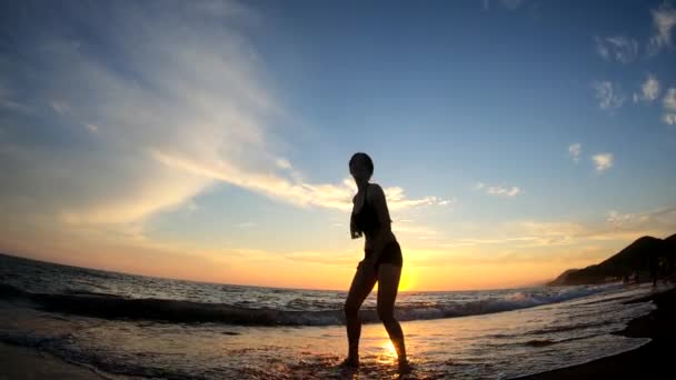 Joyeux jeune femme saute à plusieurs reprises sur fond de coucher de soleil doré à la plage de sable dans les vagues de mer. Silhouette à mouvement lent — Video